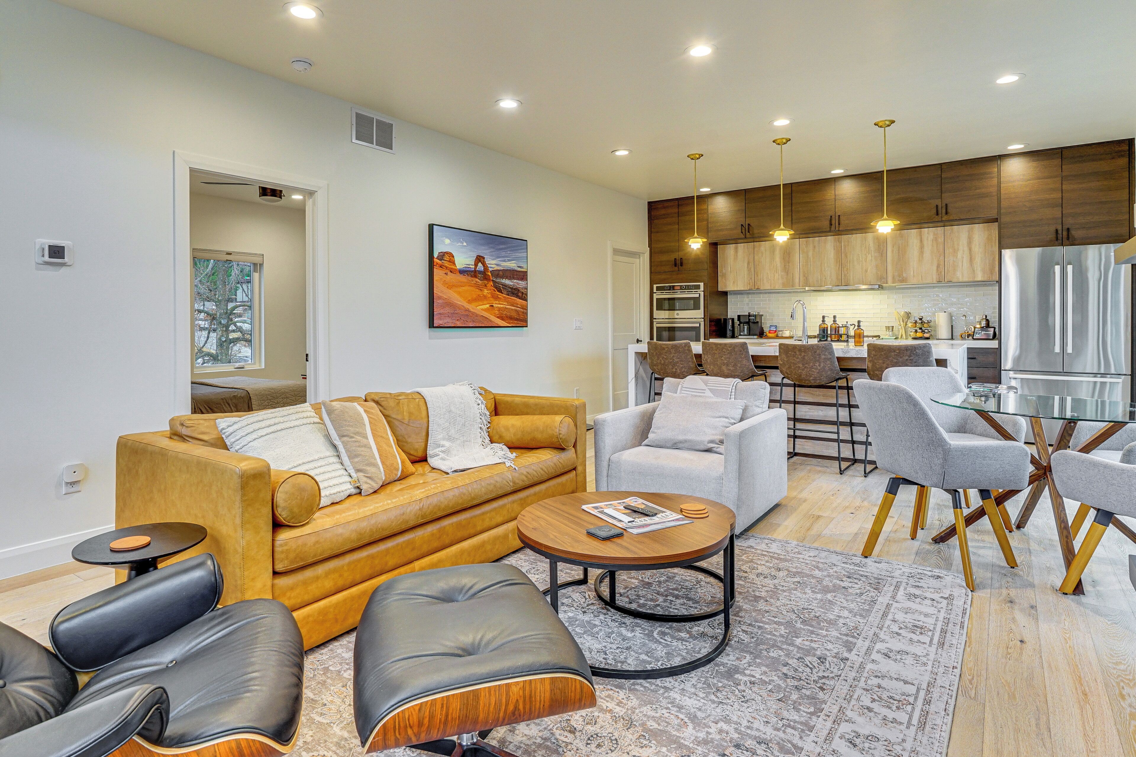 A modern living room with a sofa, armchairs, and a coffee table. There is a kitchen area with wooden cabinets and stainless steel appliances.