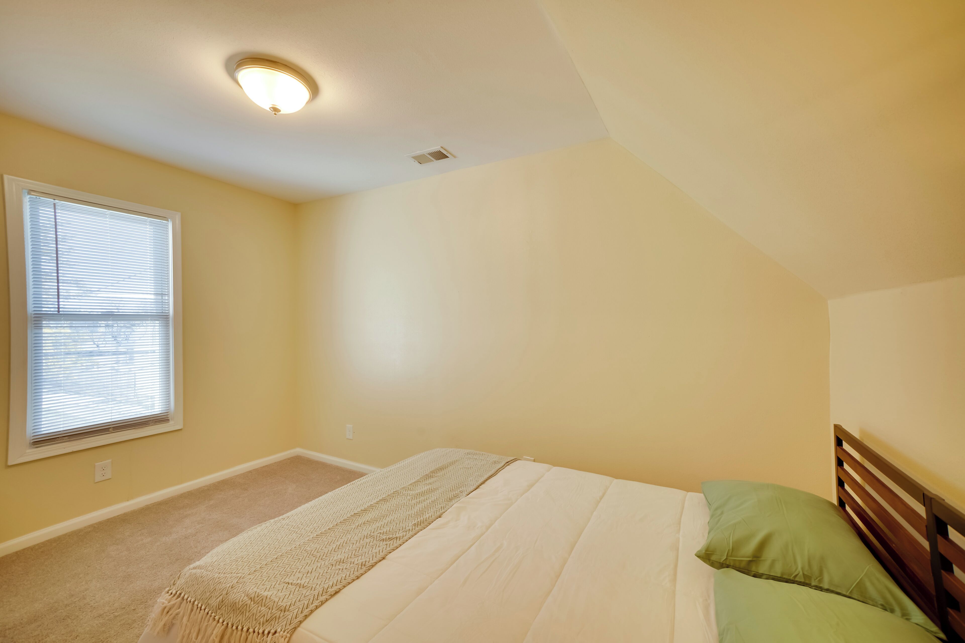 A bedroom with a single bed, a window with blinds, a ceiling light, and a carpeted floor.