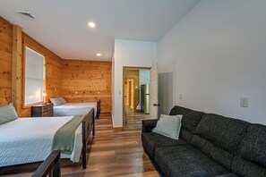 A room with two beds, a sofa, wooden walls, a staircase, and a kitchen area. - Shady Point Retreat w/ Hot Tub & Pickleball! (Killbuck)