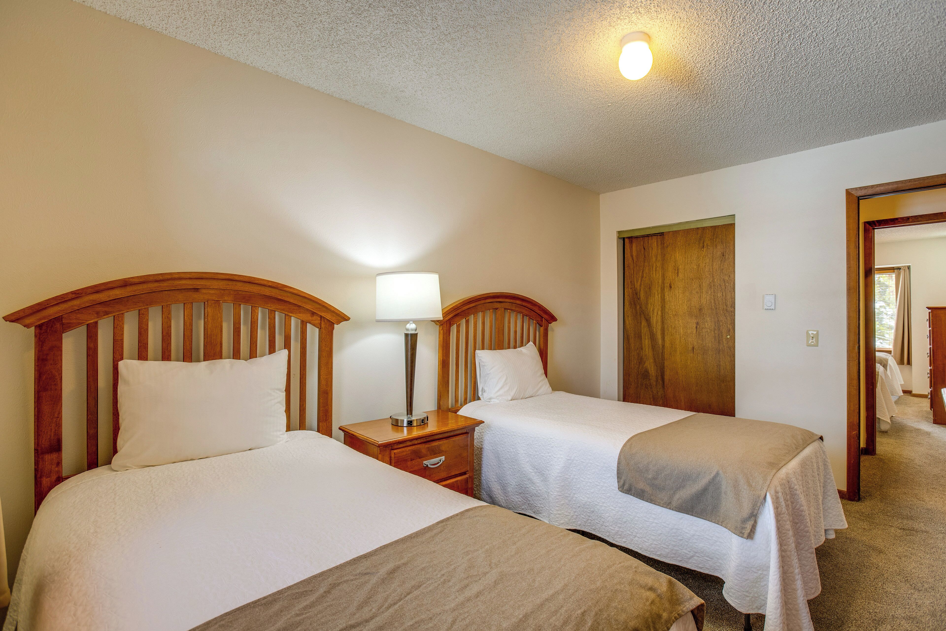 A hotel room with two beds, wooden headboards, a nightstand with a lamp, and a door leading to another room.