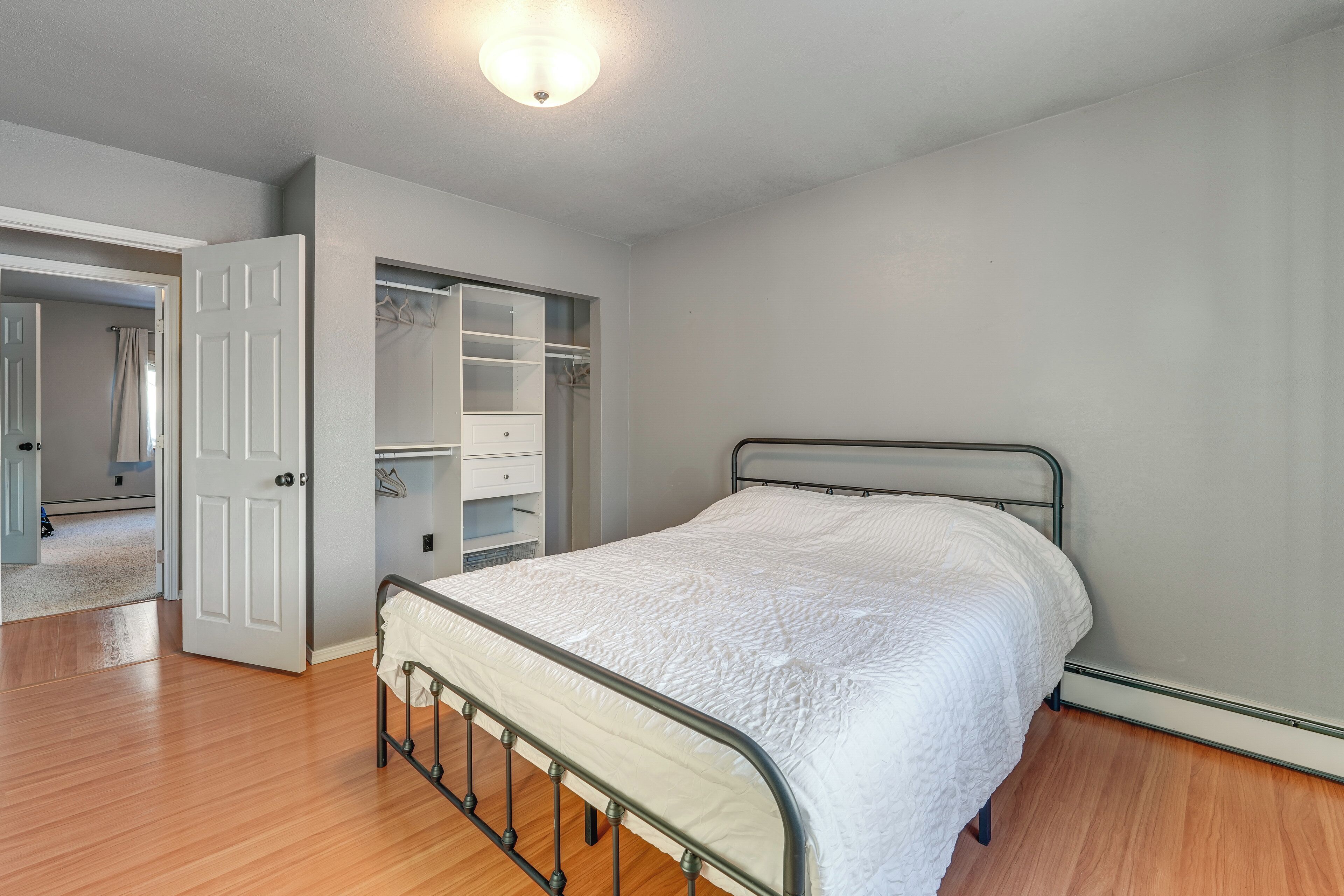 Ein Schlafzimmer mit Metallbettgestell, weißer gesteppter Bettdecke, begehbarem Kleiderschrank mit geöffneten Türen und Holzboden.
