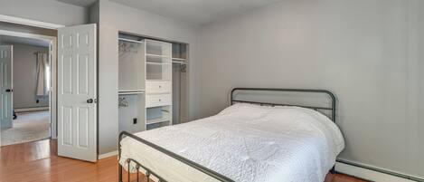Ein Schlafzimmer mit Metallbettgestell, weißer gesteppter Bettdecke, begehbarem Kleiderschrank mit geöffneten Türen und Holzboden.
