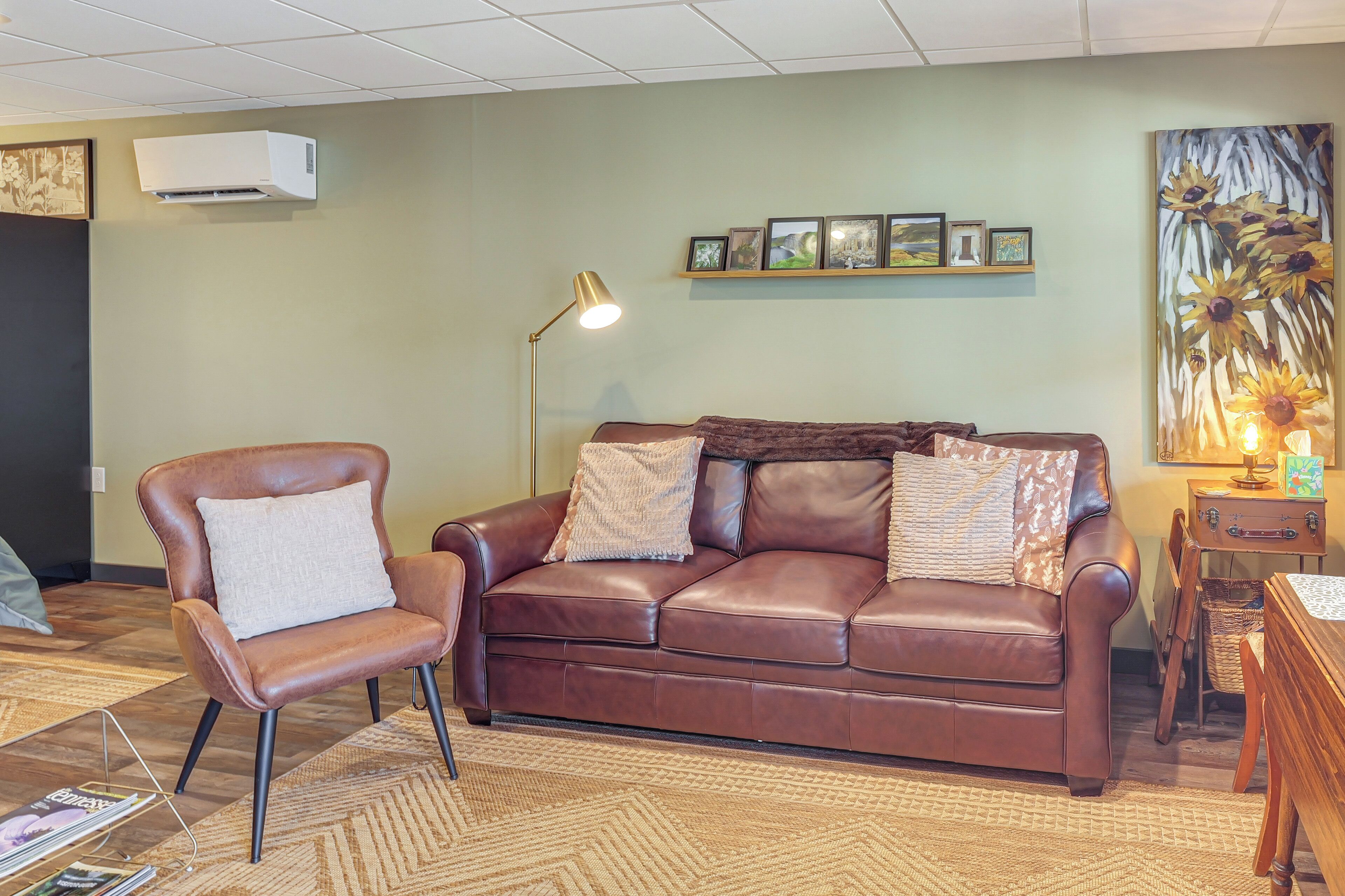 A living room with a brown leather sofa, a brown leather armchair, a floor lamp, a framed picture collection on the wall, and a painting of sunflowers.