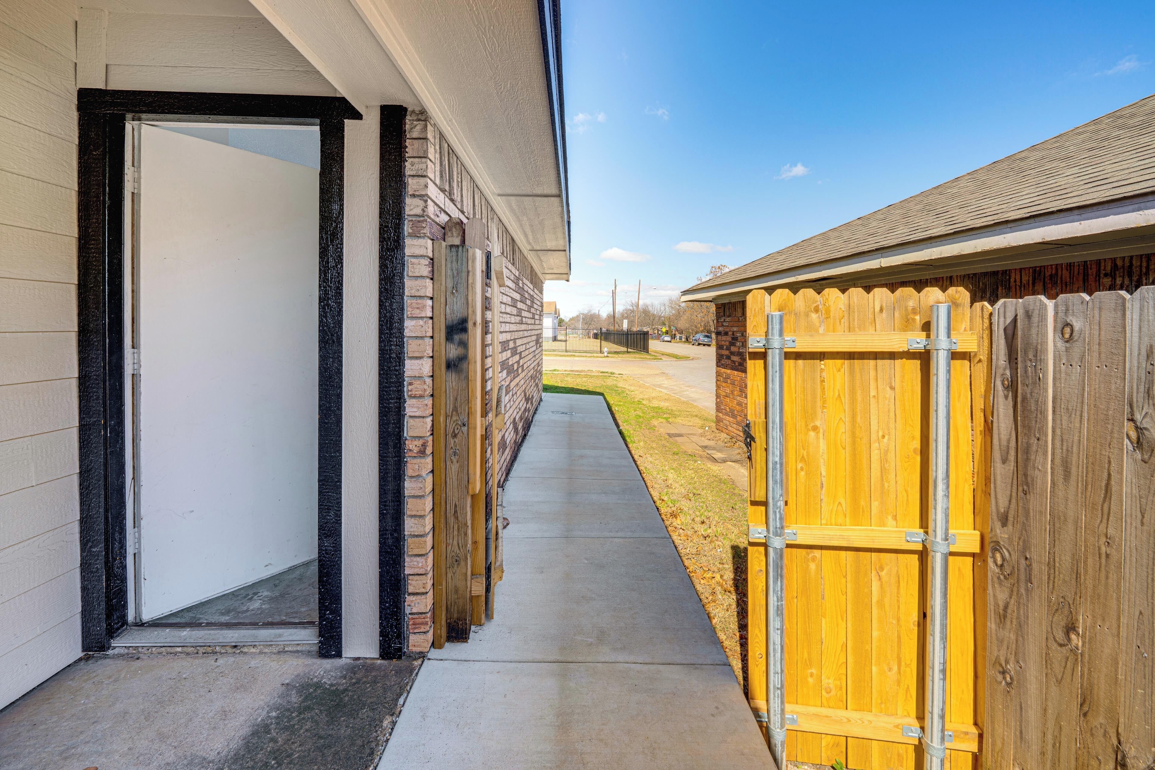 A narrow pathway leading to a building with a wooden fence on one side and a brick wall on the other.