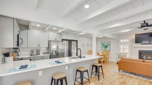 Una cocina moderna con armarios blancos, una isla central con taburetes y una zona de comedor con sofá y chimenea.