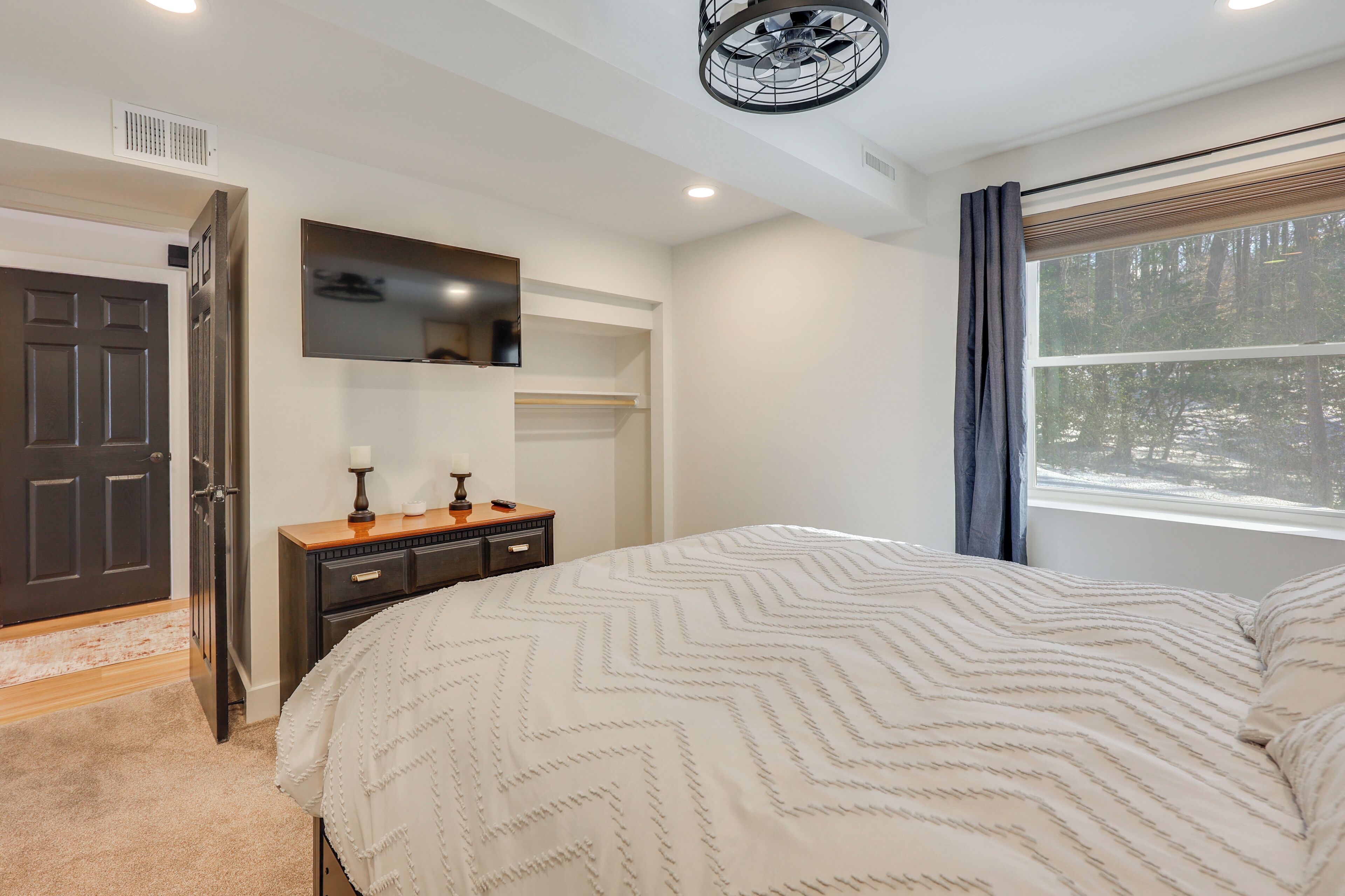 Une chambre à coucher avec un lit, une commode, un miroir et une fenêtre donnant sur des arbres.