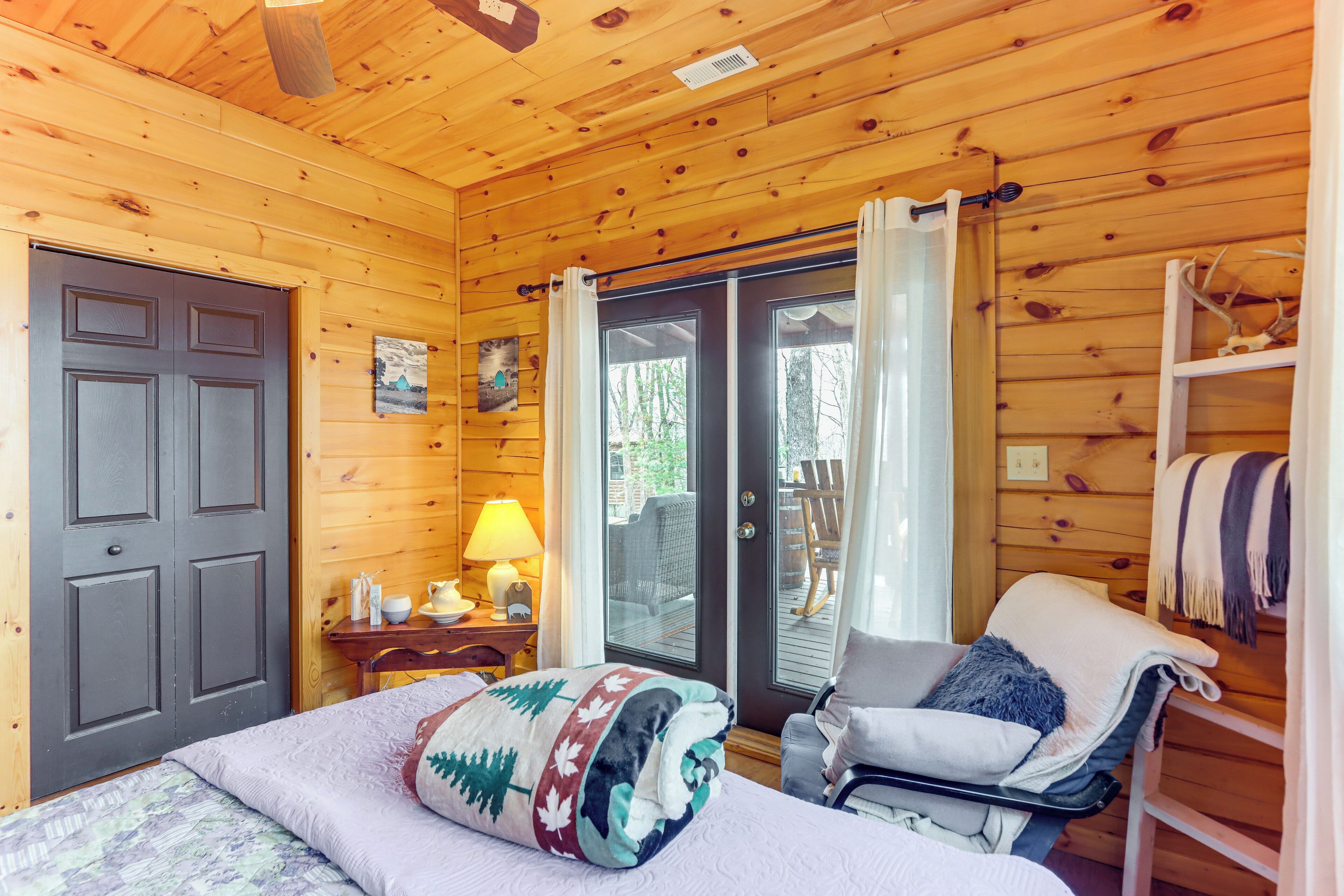 A cozy wooden cabin room with a bed, a chair, a table with a lamp, and a sliding glass door leading to an outdoor area.