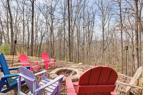 Un’area boscosa con un focolare e diverse poltrone Adirondack di vari colori.