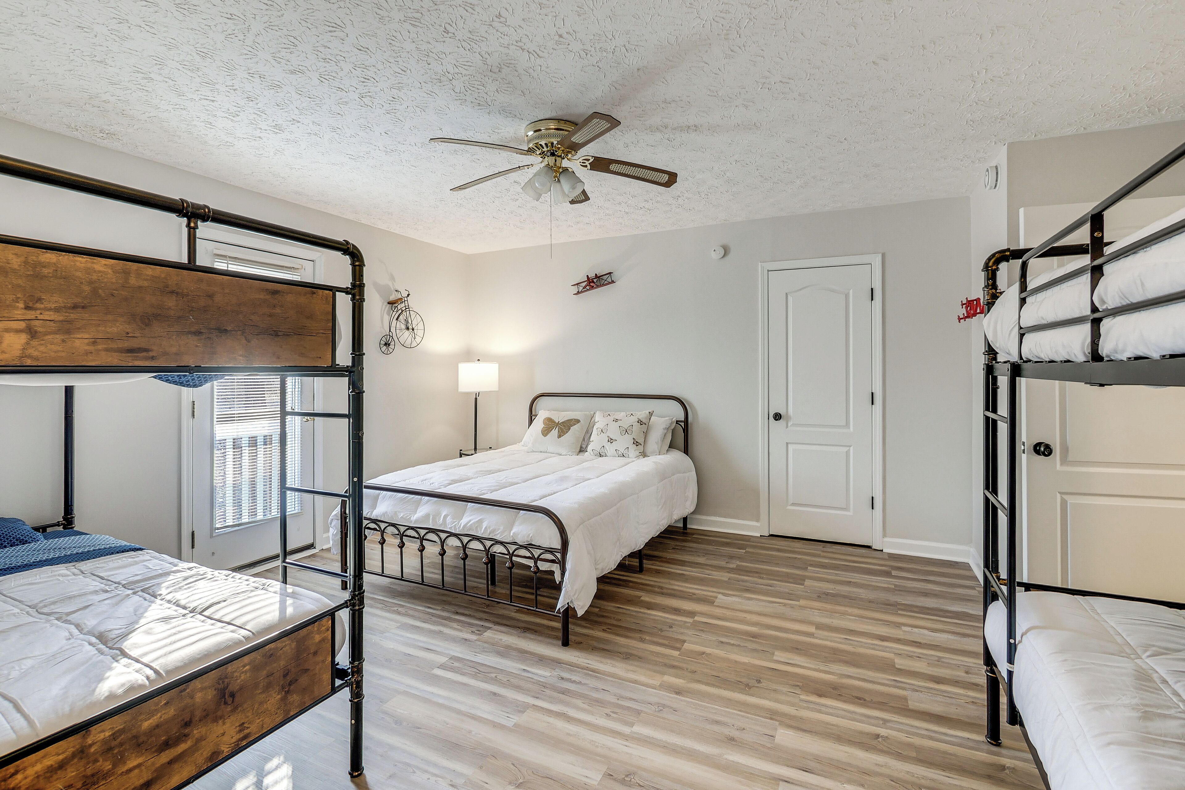 A dormitory room with bunk beds, a ceiling fan, a bedside lamp, and a door.
