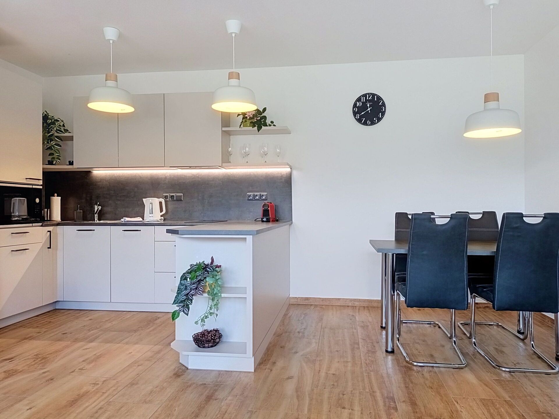 A modern kitchen with white cabinets, a central island with plants, a dining area with black chairs, and a wall clock.