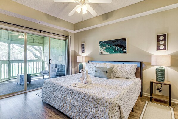 A bedroom with a large bed, a ceiling fan, a painting, and a view through a sliding glass door.