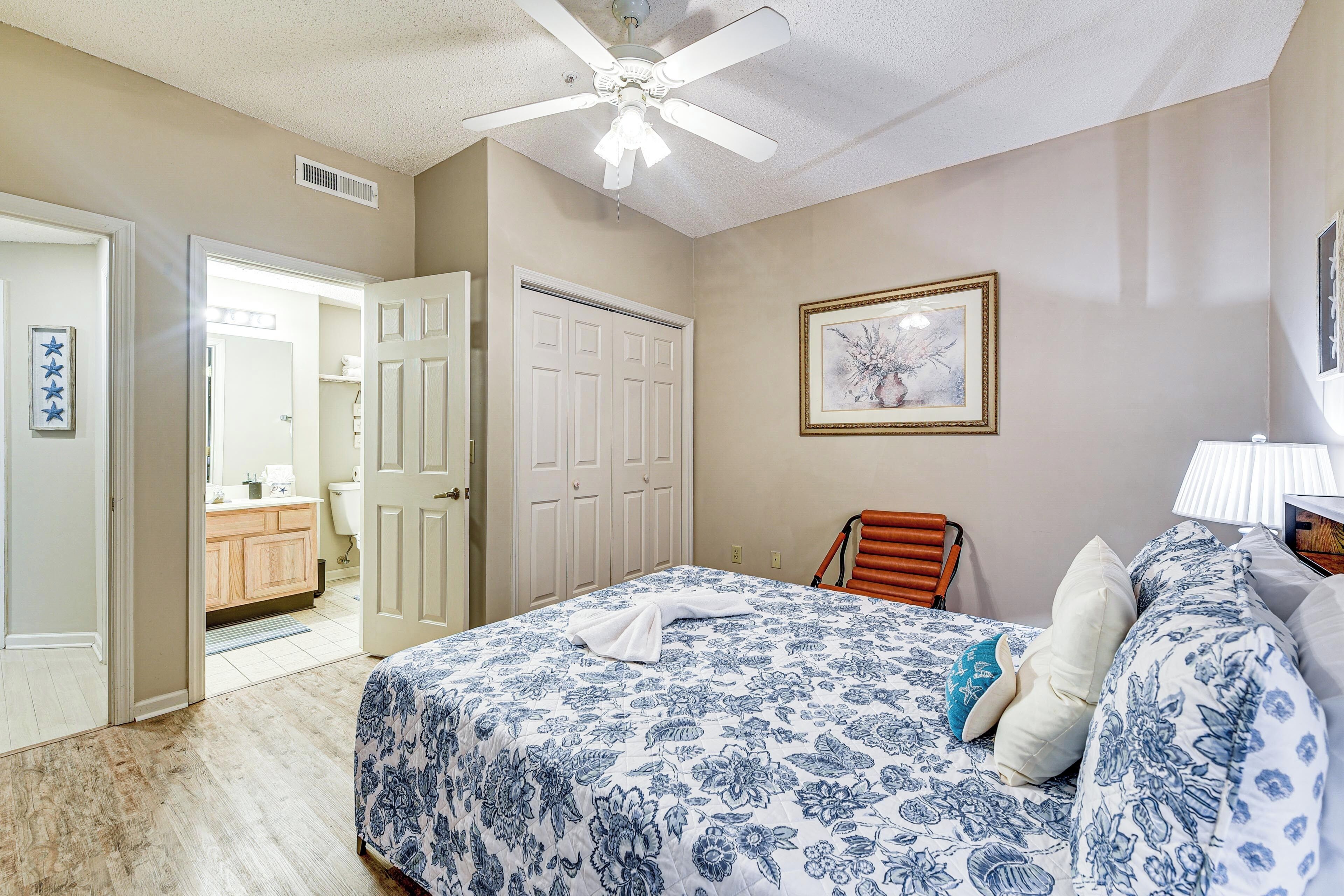 A bedroom with a bed, a ceiling fan, a door, a framed picture, and a nightstand with a lamp.