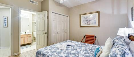 A bedroom with a bed, a ceiling fan, a door, a framed picture, and a nightstand with a lamp.