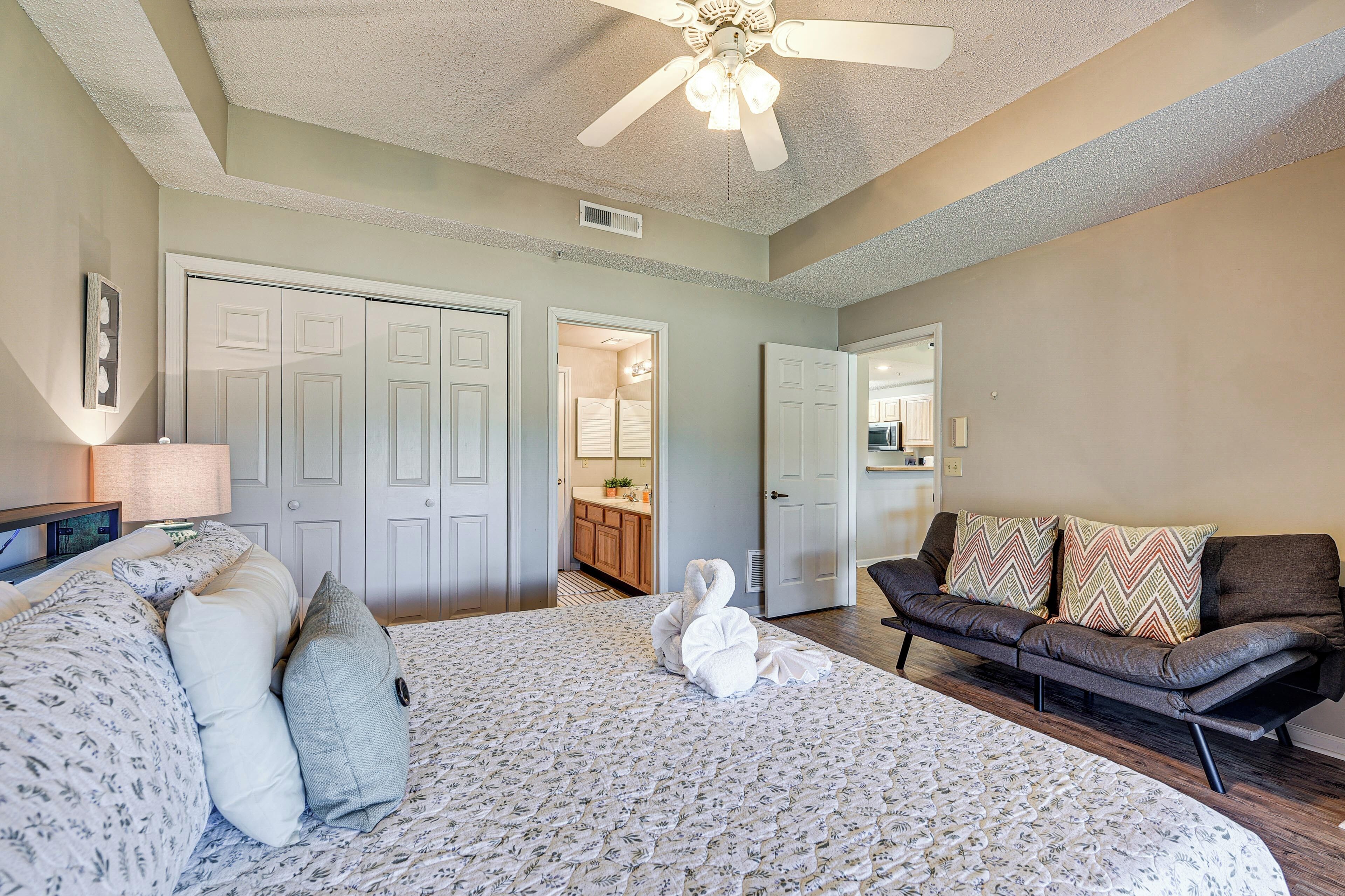 A bedroom with a bed, a sofa, a ceiling fan, and a door leading to a bathroom.
