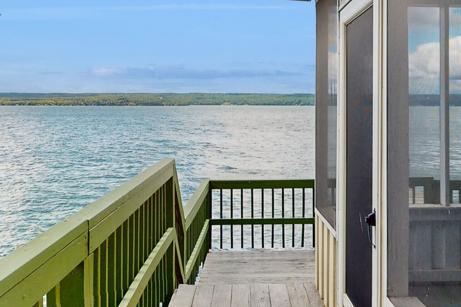 Une terrasse en bois avec une rambarde verte mène à un bâtiment doté d’une porte en verre, qui offre une vue sur une étendue d’eau calme et une côte lointaine.