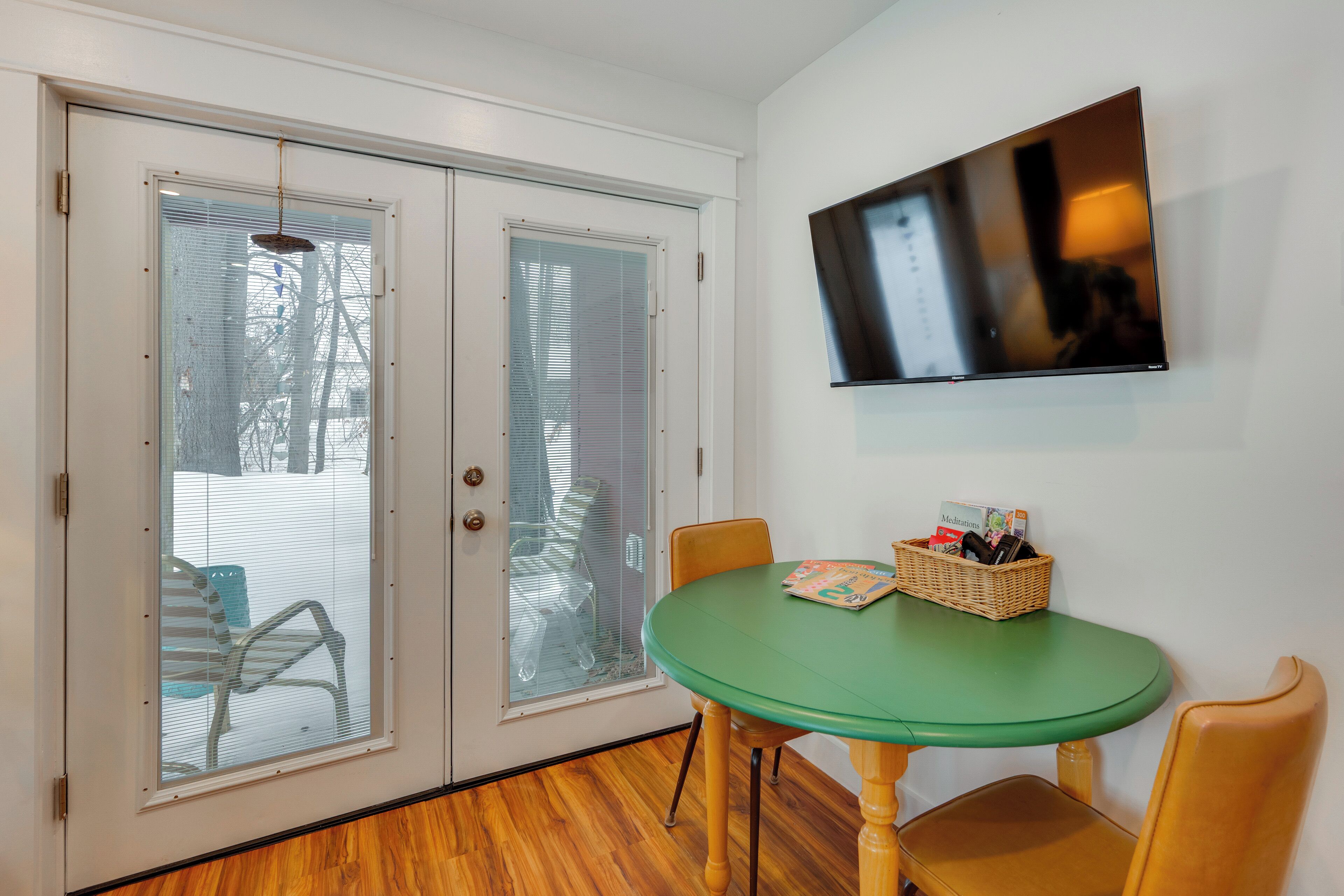 A room with a green table, two chairs, a wicker basket with magazines, and a TV on the wall.