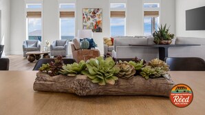 Un salon spacieux avec un grand canapé, deux fauteuils, une table basse et une cheminée. De grandes fenêtres sont équipées de stores, et le salon est doté d’un ventilateur de plafond et d’un téléviseur à écran plat fixé au mur.