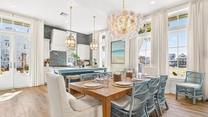 A spacious dining area with a wooden table and wicker chairs, a kitchen with white cabinets, and large windows offering a view of the city. - Popp’s Place | Preppy Coastal Retreat Steps from Pool and Lake @CS South (Port Aransas)