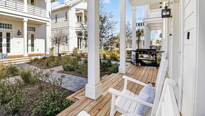 A covered porch with white columns and a wooden deck, surrounded by a garden with various plants and a barbecue grill. - Popp’s Place | Preppy Coastal Retreat Steps from Pool and Lake @CS South (Port Aransas)