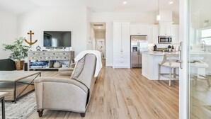 A spacious living room with a beige sofa, a TV, and a wooden coffee table. A kitchen with white cabinets and a microwave is visible in the background. - Blessing By The Bay | Community Pool | Sleeps 8 (Galveston)