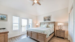 A bedroom with a wooden bed, two bedside tables, a ceiling fan, and a view of the outside through a door. - Blessing By The Bay | Community Pool | Sleeps 8 (Galveston)