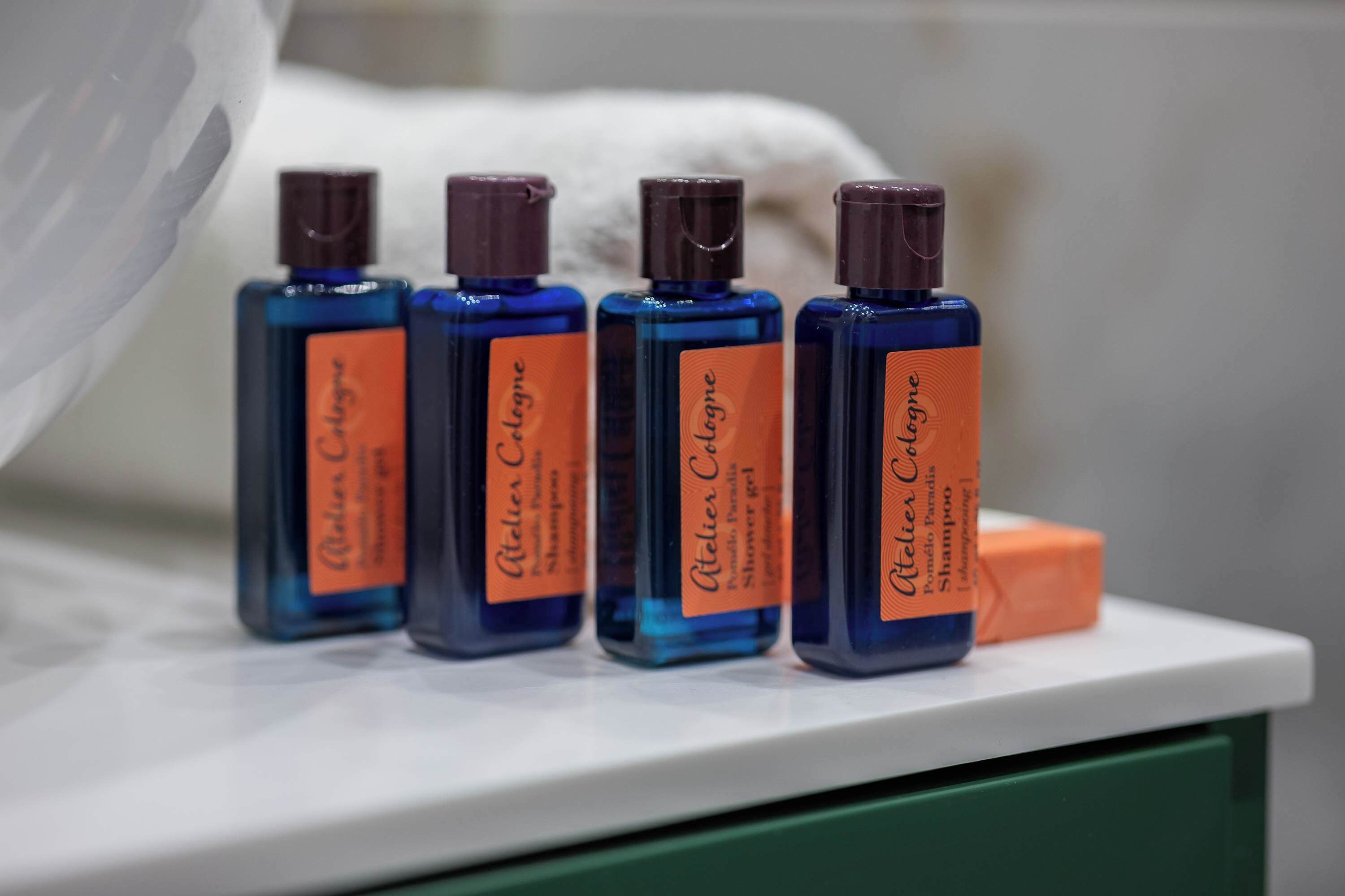 Four bottles of Ateliers Cologne shampoo and conditioner on a white shelf.