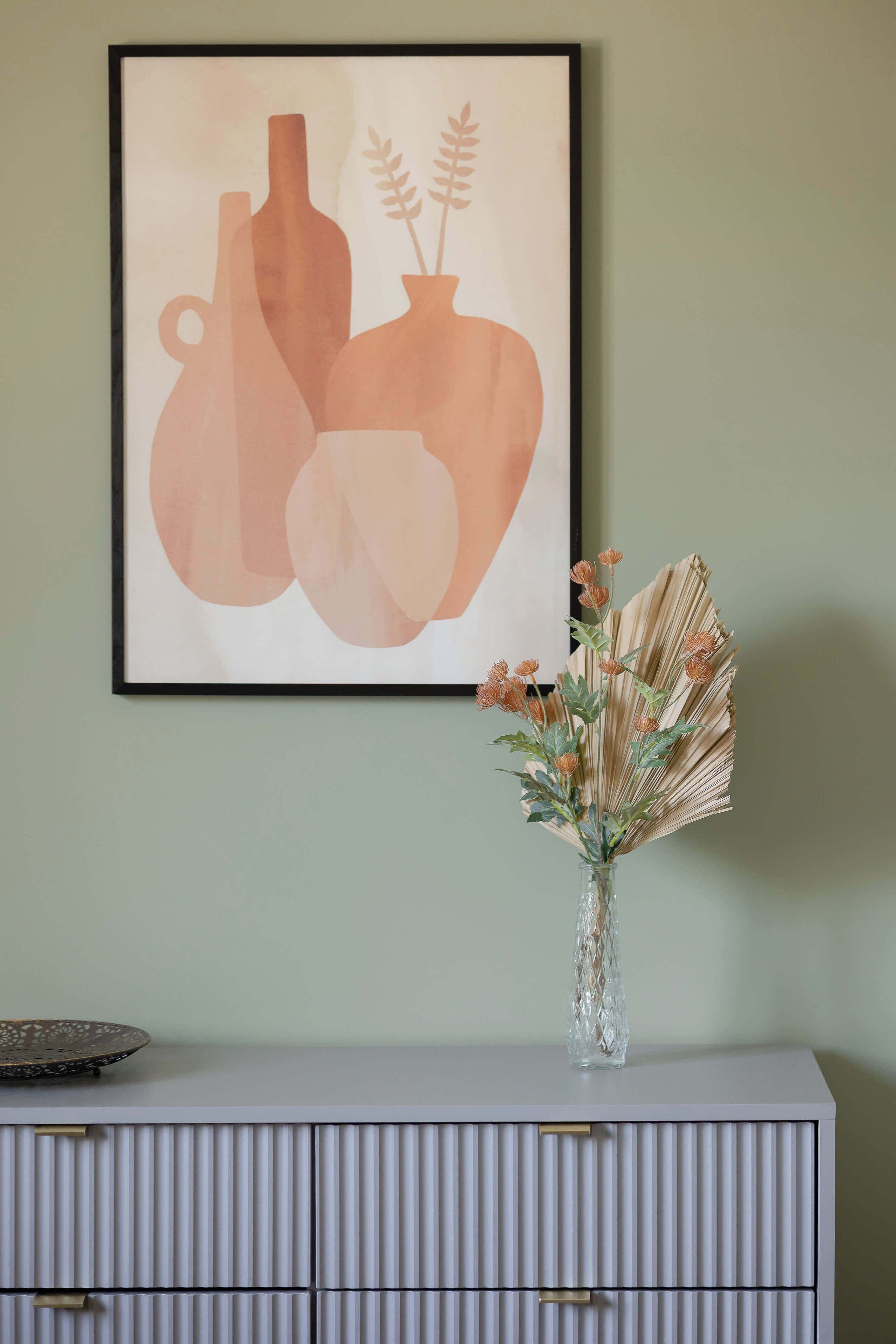 Une œuvre d’art encadrée représentant des vases abstraits et un vase contenant des fleurs séchées, le tout posé sur un meuble blanc.