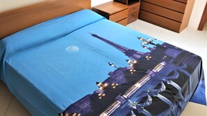Una cama con colcha azul, cabecera de madera, mesita de noche de madera y cómoda de madera.