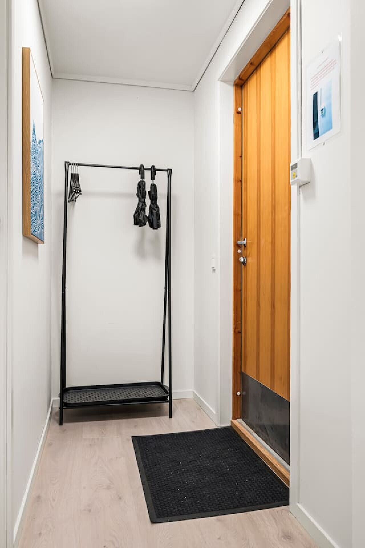 Un couloir étroit avec un banc en métal noir, une porte en bois et un paillasson noir à l’entrée.