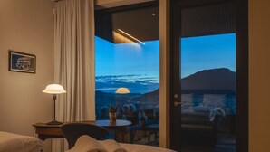 Una camera d'albergo con un letto, una scrivania, una lampada e vista su una città e montagne.
