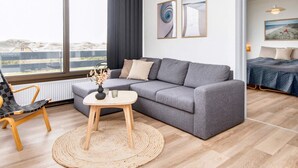 Ein modernes Wohnzimmer mit einem grauen Sofa, einem runden Couchtisch aus Holz und einem gewebten Teppich. Durch große Fenster bietet sich ein Blick ins Freie, und der Raum zeichnet sich durch ein minimalistisches Design mit einem gerahmten Bild an der Wand aus.