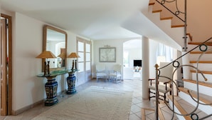 A spacious hallway with a glass console table, two lamps, and a large mirror. A white rug covers the tiled floor, leading to a staircase with a metal railing.