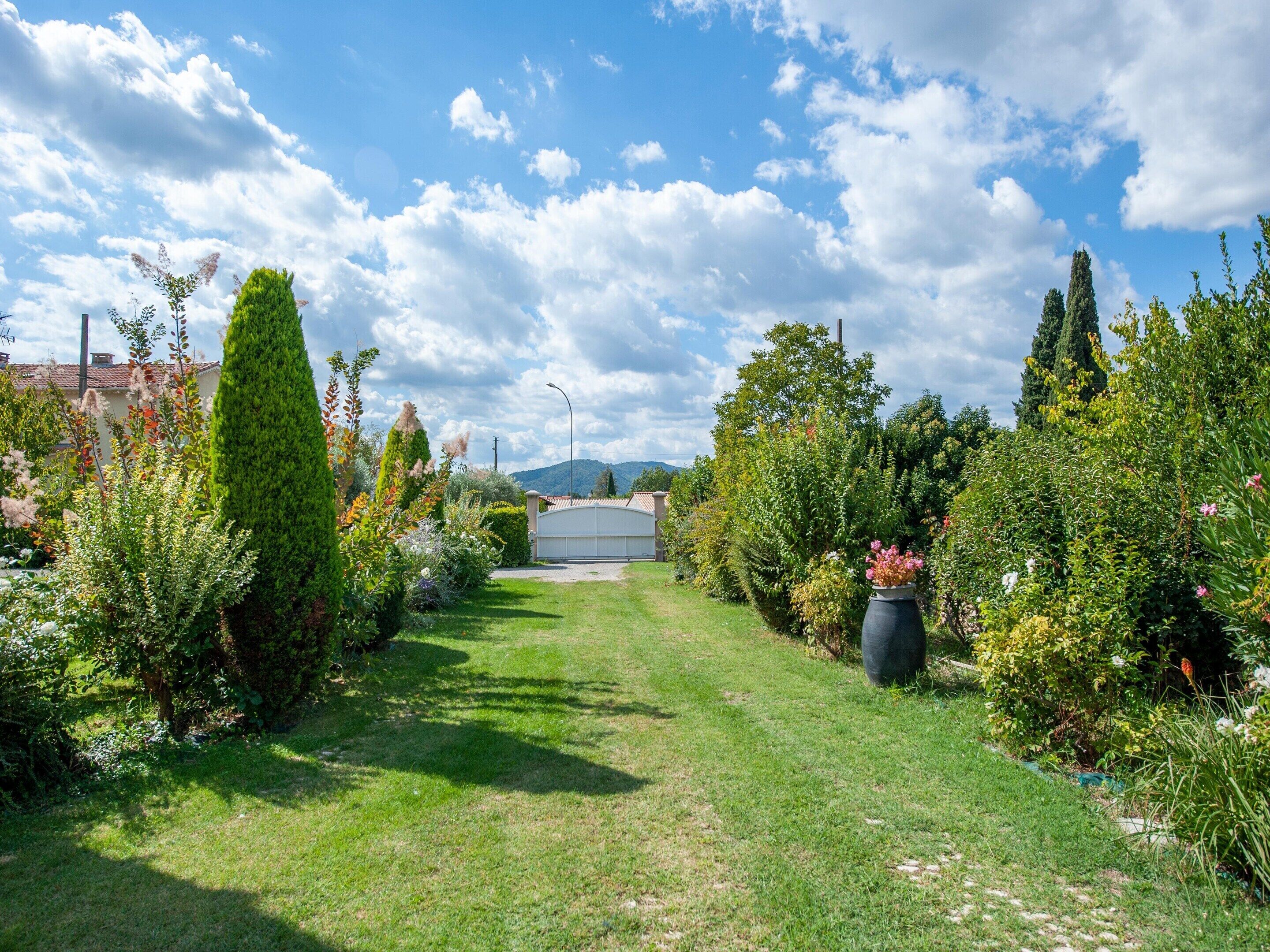 A well-maintained garden with a variety of plants and trees, a clear sky, and a pathway leading to a building.