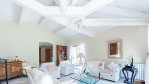 A spacious living room with white furniture, a glass coffee table, and a large bookshelf.