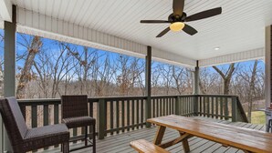 A covered outdoor deck with a wooden table and two wicker chairs, a ceiling fan, and a view of leafless trees. - The Allendale Overlook (Bella Vista)