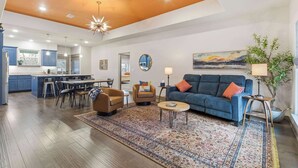 A spacious living room with a blue sofa, a coffee table, and a patterned rug. There is a kitchen area with blue cabinets and a dining table. - The Allendale Overlook (Bella Vista)