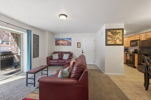 A living room with a leather sofa, a bench, and a sliding glass door leading to an outdoor area. There is a kitchen with wooden cabinets and appliances. - New Listing! Adventure Ready | Near Arches | Hot Tub + Pool | Views (Moab)