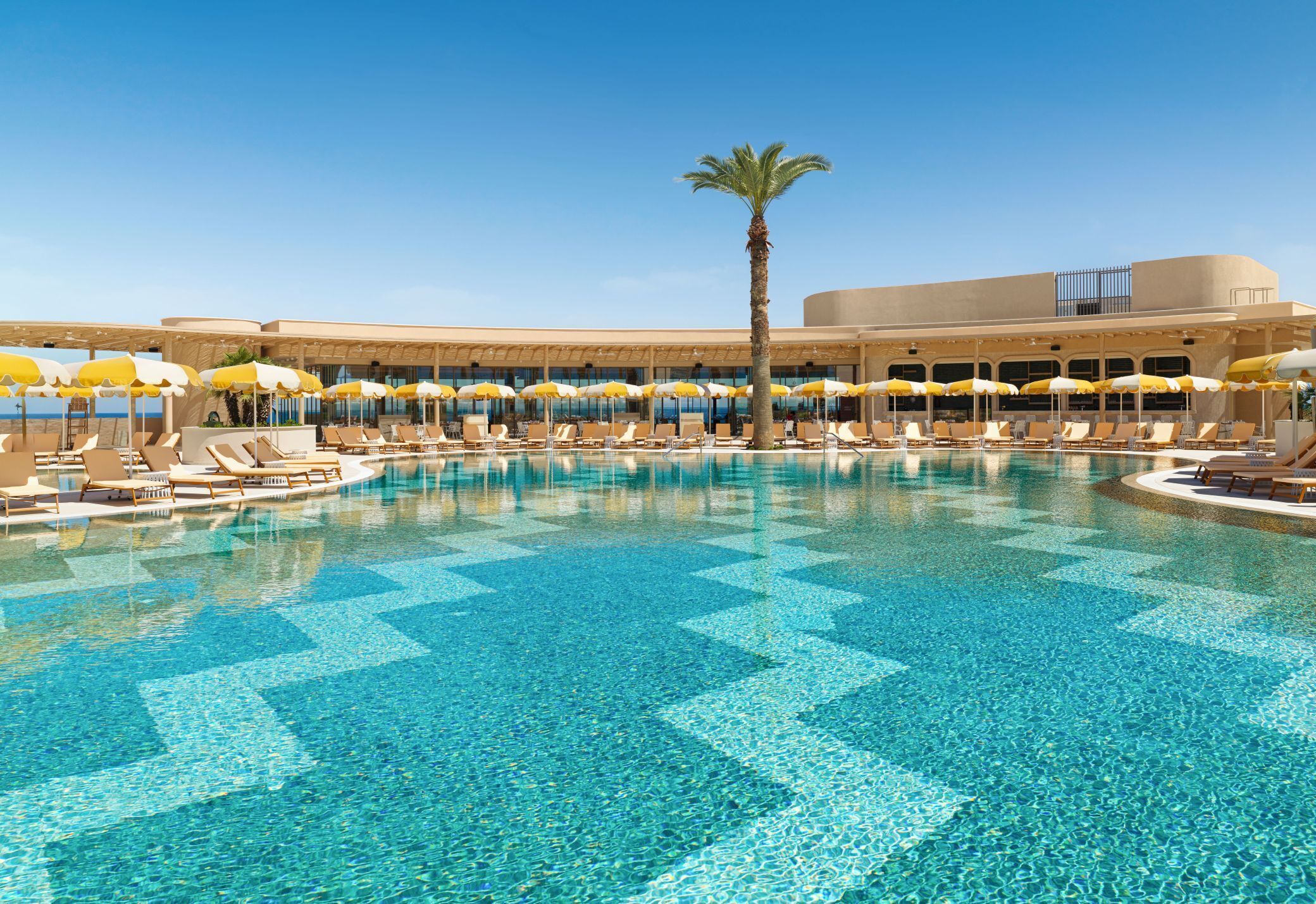 A pool with clear blue water, surrounded by lounge chairs and umbrellas, and a palm tree.