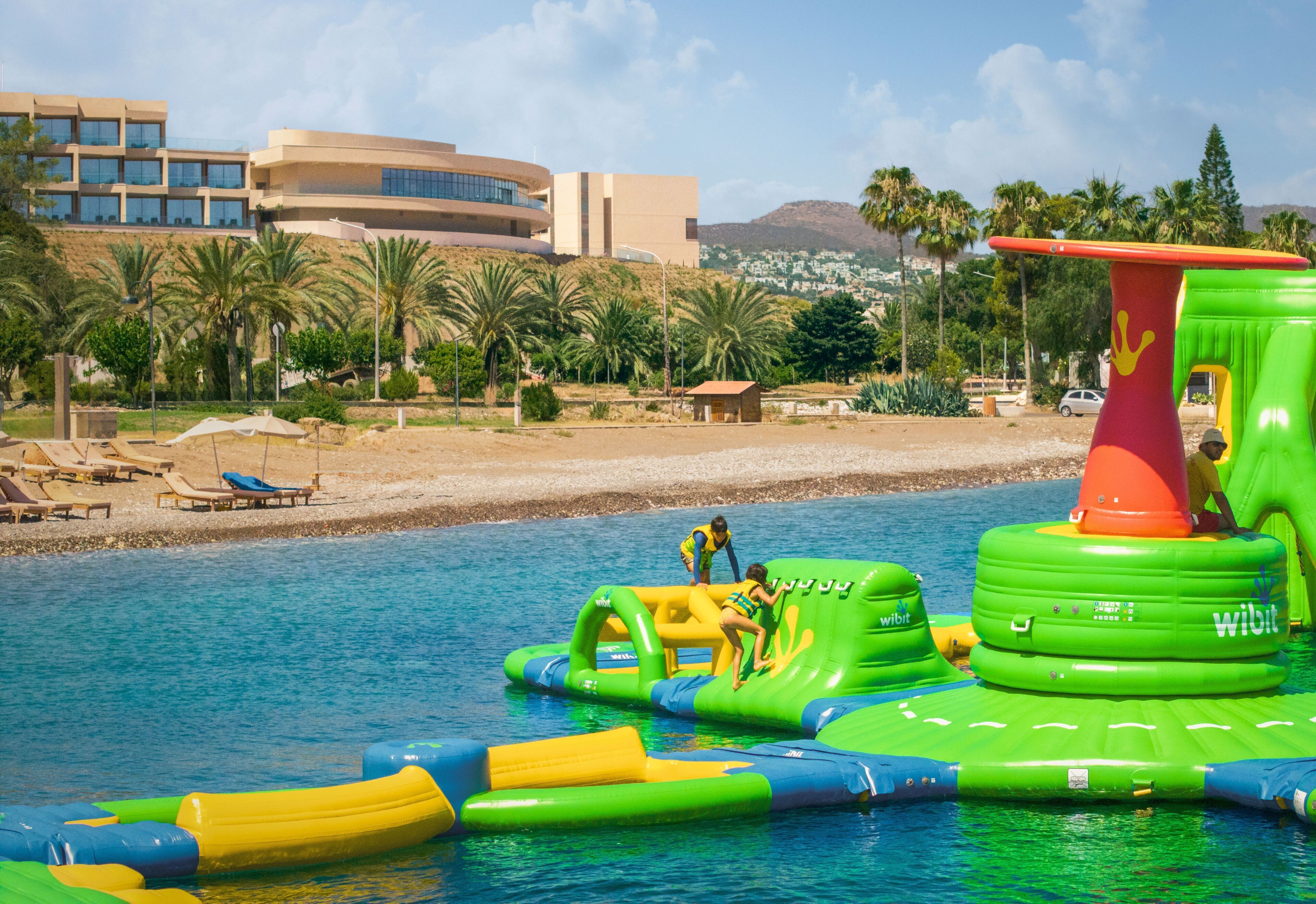 An inflatable water park with slides and a pool, surrounded by palm trees and a building in the background.