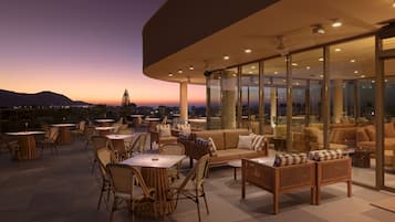 An outdoor seating area with tables and chairs, a sofa, and a view of the sunset over the mountains.