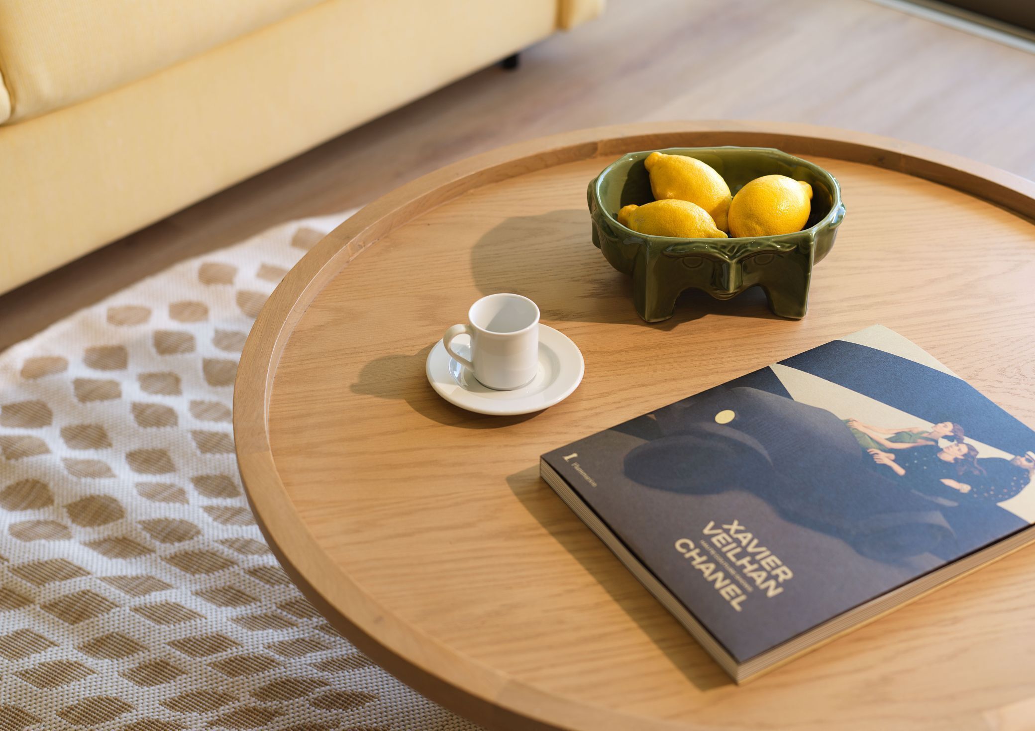 Una mesa de madera redonda con una taza y platillo blancos, un bol de limones y un libro titulado 'Xavier Veilhan Chanel'.