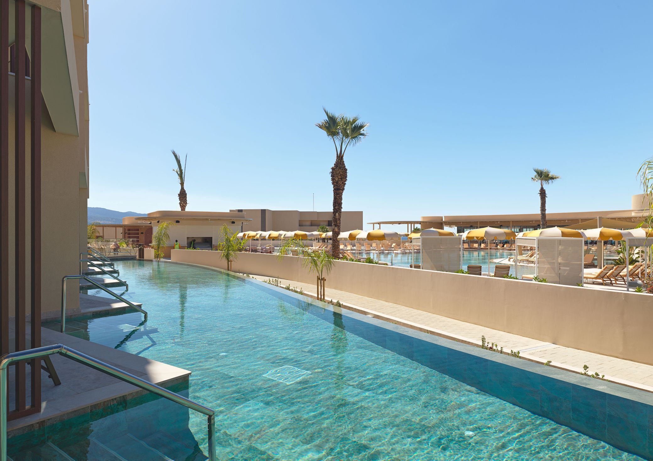 A clear swimming pool with sun loungers and umbrellas, surrounded by palm trees and a clear sky.