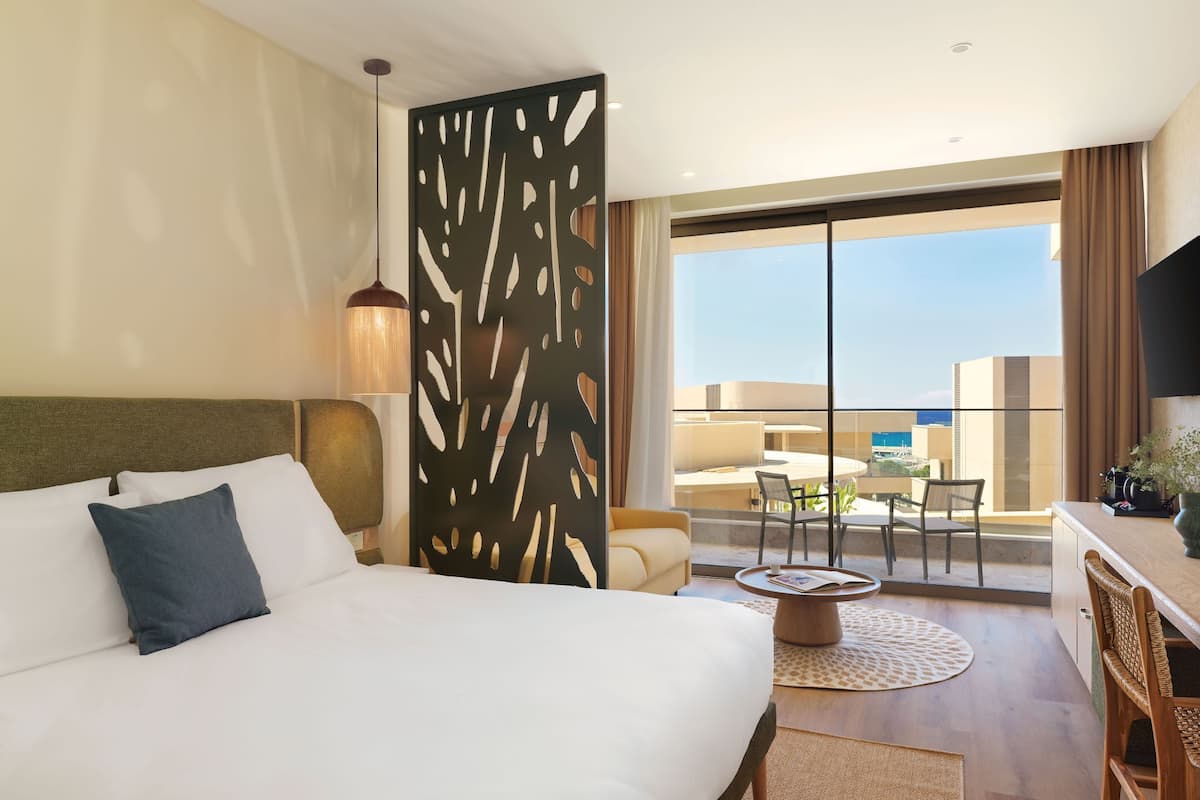 Habitación de hotel con cama, escritorio, silla, mesa de comedor y balcón con vistas.