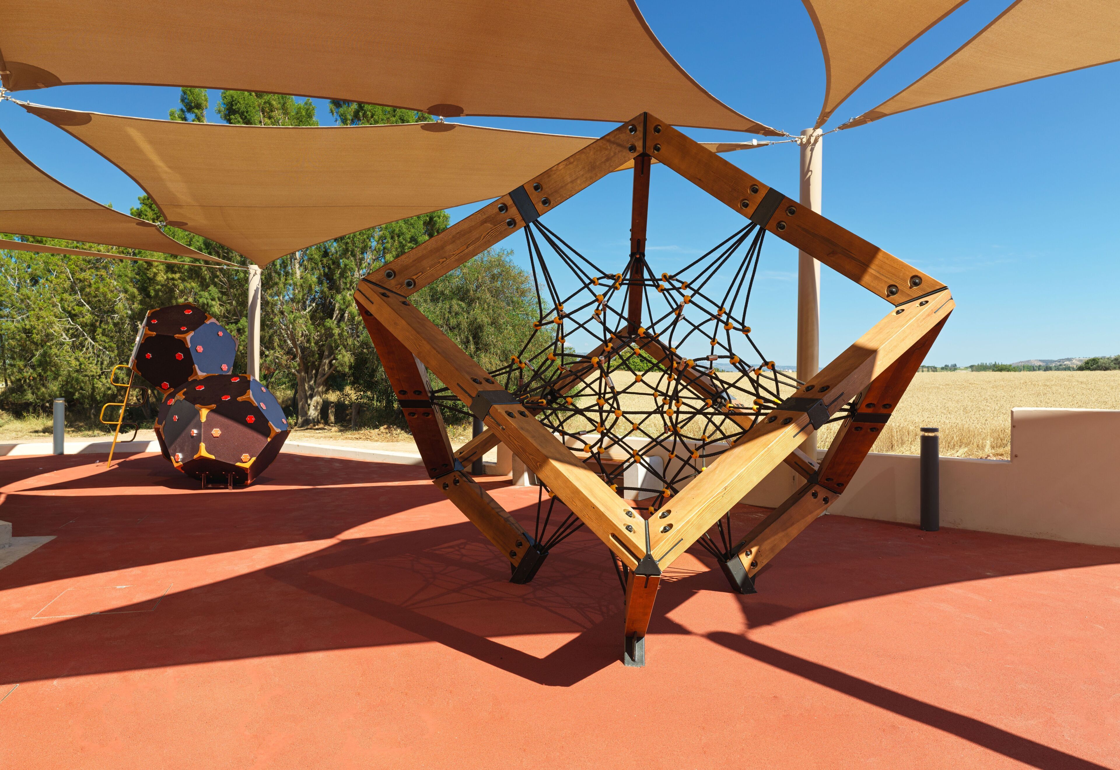 A geometric wooden structure with a network of ropes, set on a red surface with a clear sky in the background.