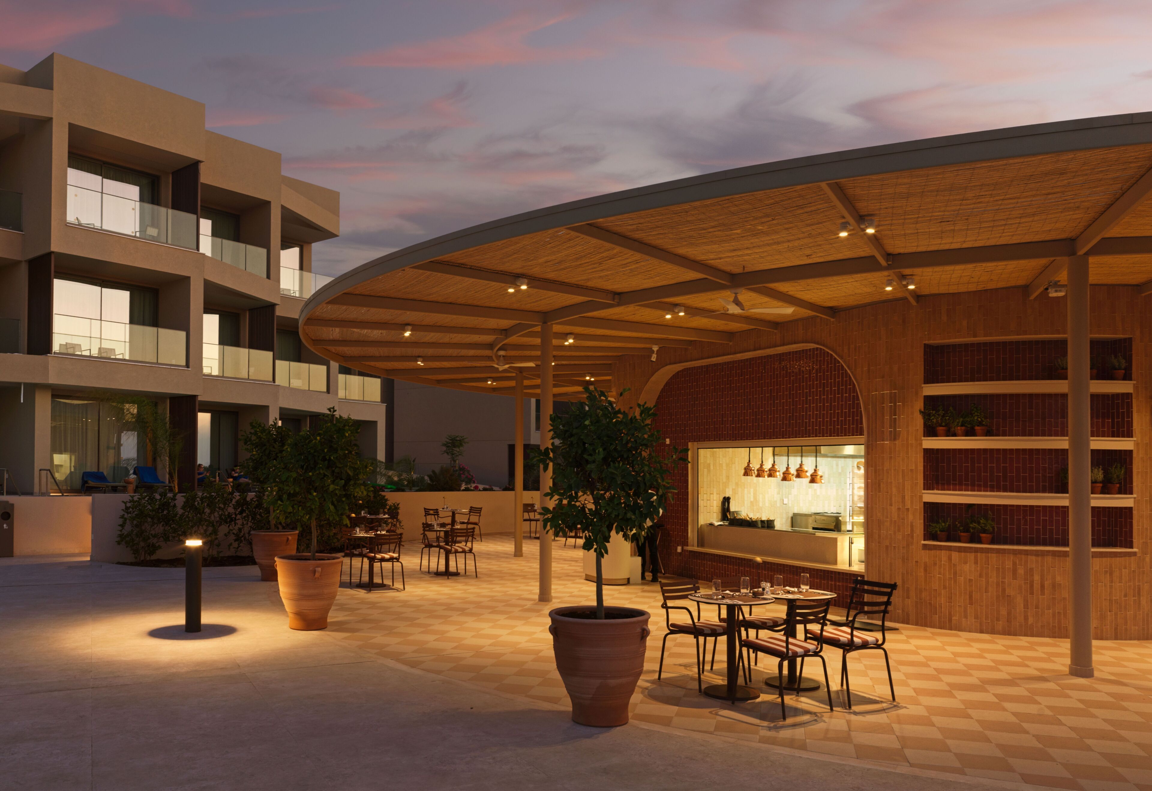 A modern outdoor dining area with multiple tables and chairs, surrounded by large potted plants and a covered seating area with a wooden ceiling.