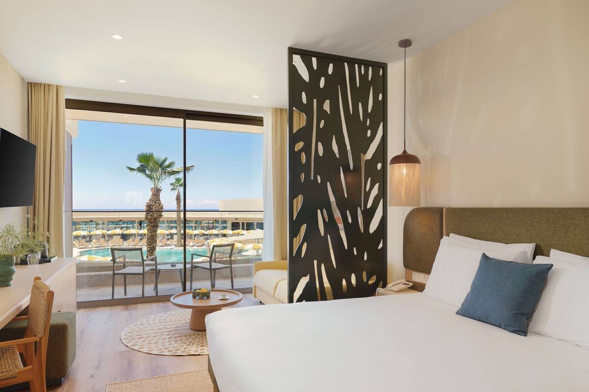 Habitación de hotel con cama, escritorio, silla, mesa redonda, vista a una piscina y palmeras, y un separador decorativo.