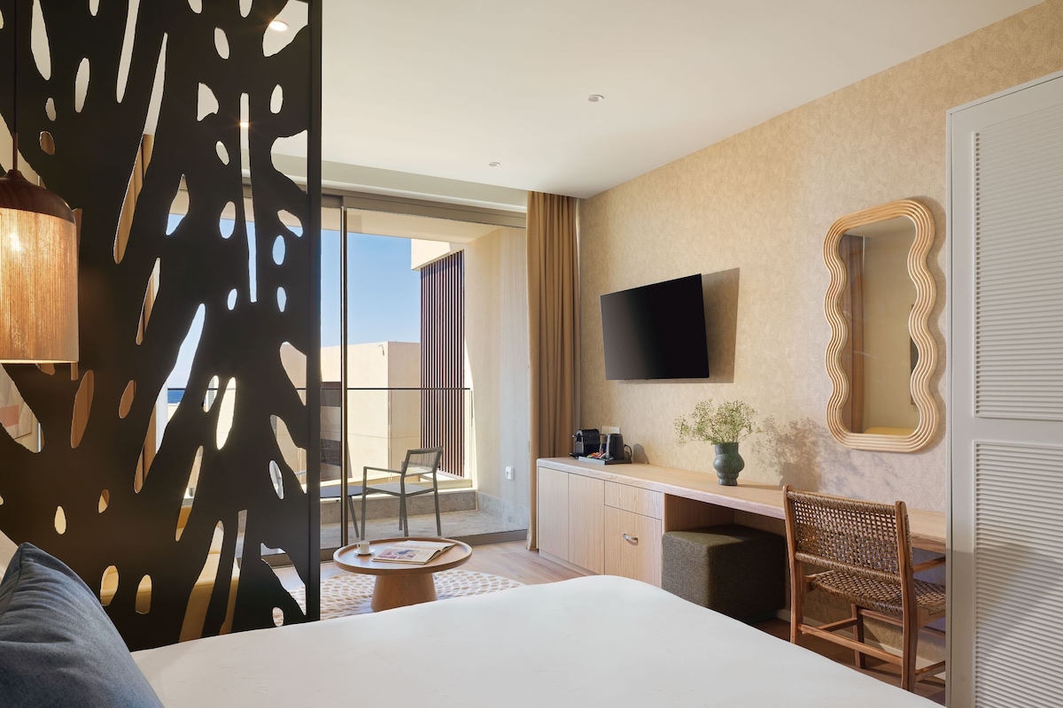 A hotel room with a bed, a desk, a chair, a TV, and a balcony with a view.