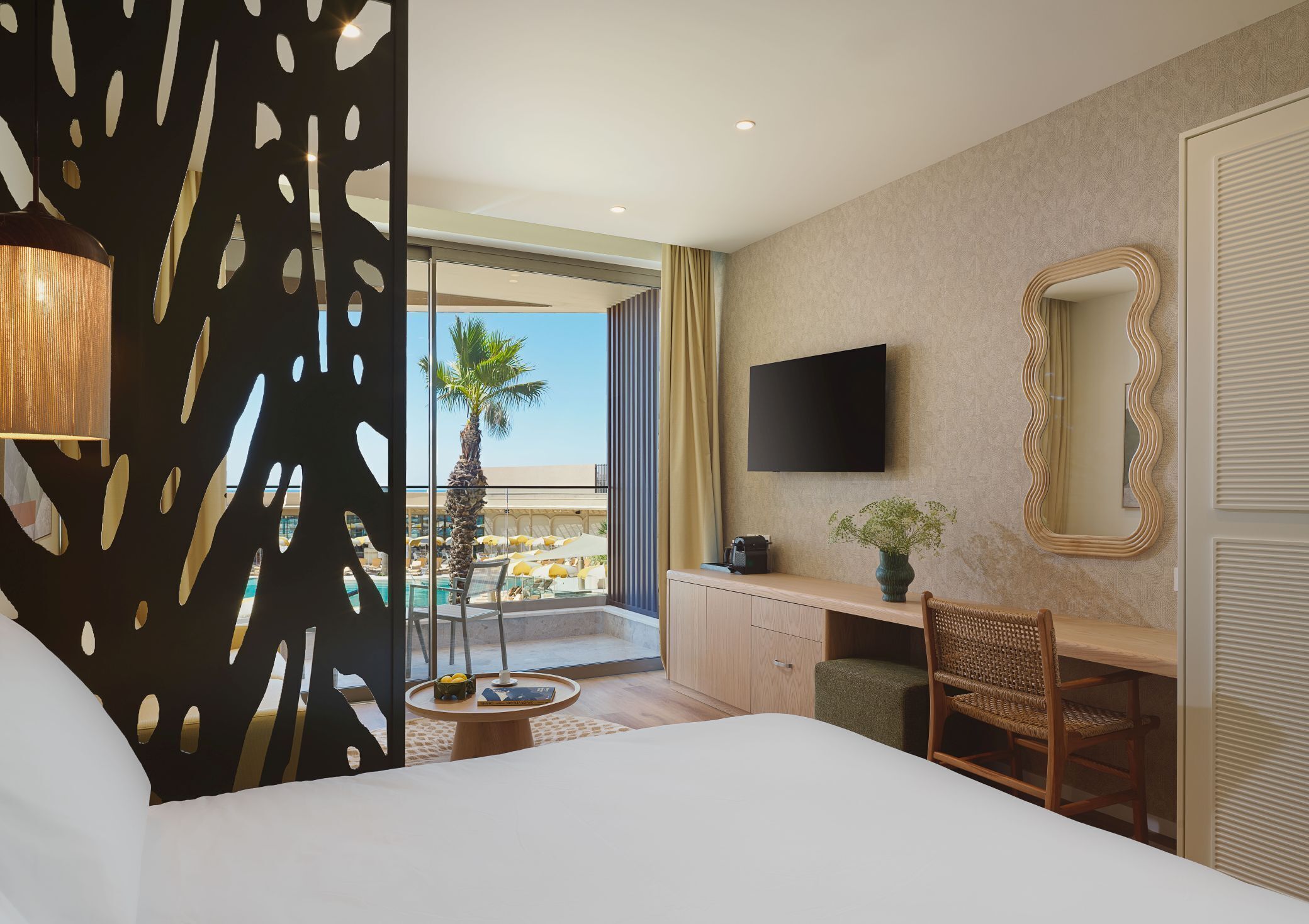 Habitación de hotel con cama, escritorio, silla, televisor, espejo y balcón con piscina y palmeras.