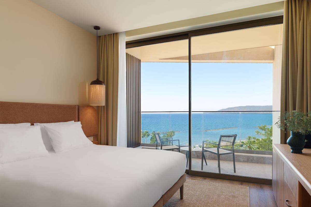 A hotel room with a bed, a nightstand, a lamp, a view of the sea, and a balcony with chairs.