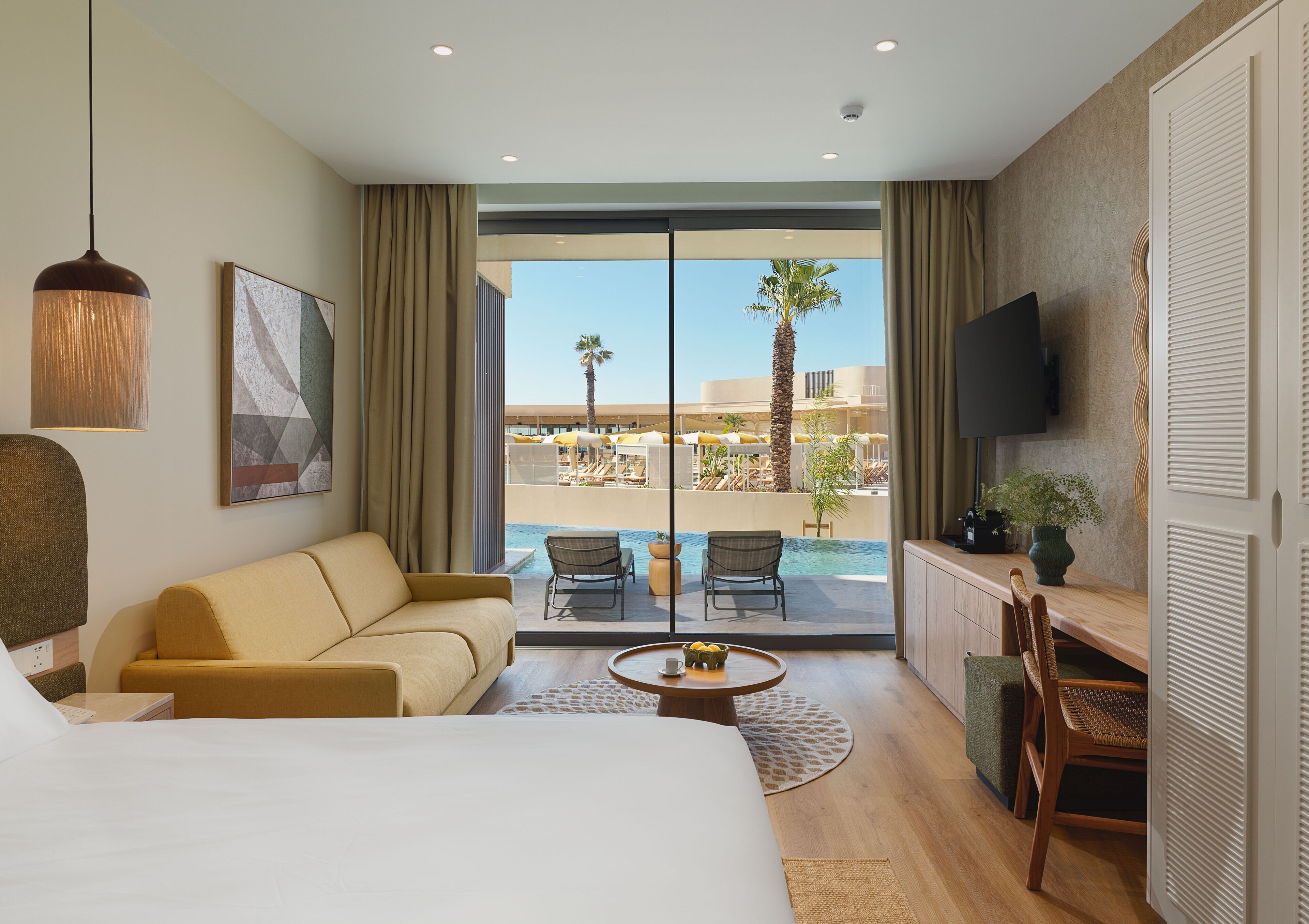 Ein Hotelzimmer mit Bett, Sofa, Esstisch, Fernseher und Blick auf einen Pool und Palmen.